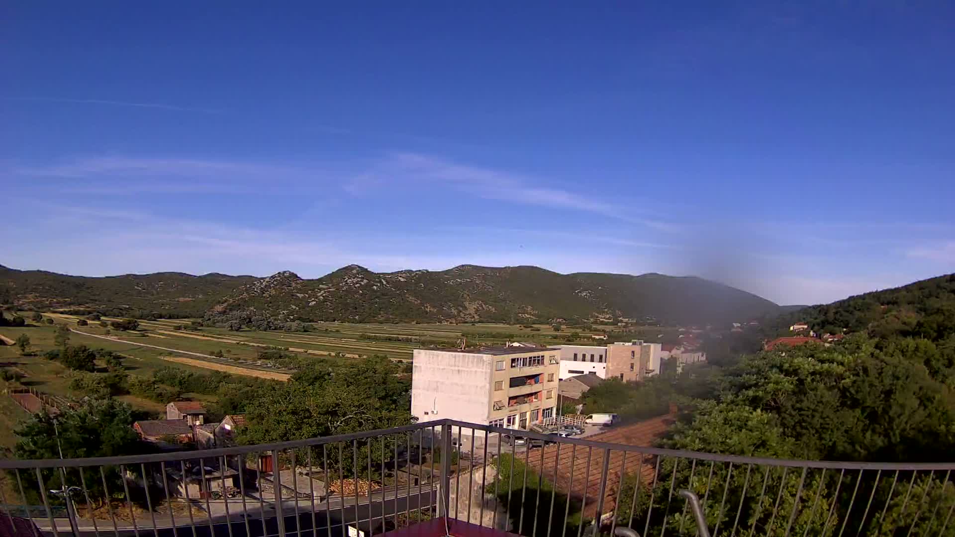A clear, sunny day reveals a valley of farmland bordered by low mountains, with buildings and trees visible in the foreground from an elevated viewpoint.
