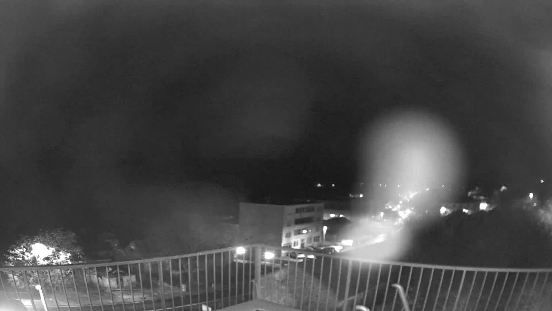 A monochromatic, blurry nighttime outdoor scene shows a town viewed from a balcony, with buildings and scattered lights partially visible through heavy fog or mist, and a large light flare dominating the right side.