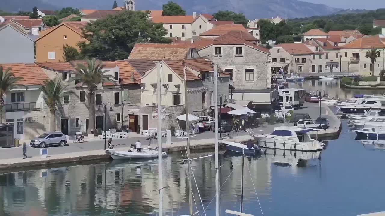 A waterfront town with stone buildings, palm trees, docked boats, and cars parked along a canal on a sunny day.