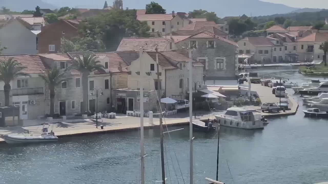 A waterfront village with stone buildings, palm trees, boats moored in a calm harbor, and several cars parked nearby on a sunny day.