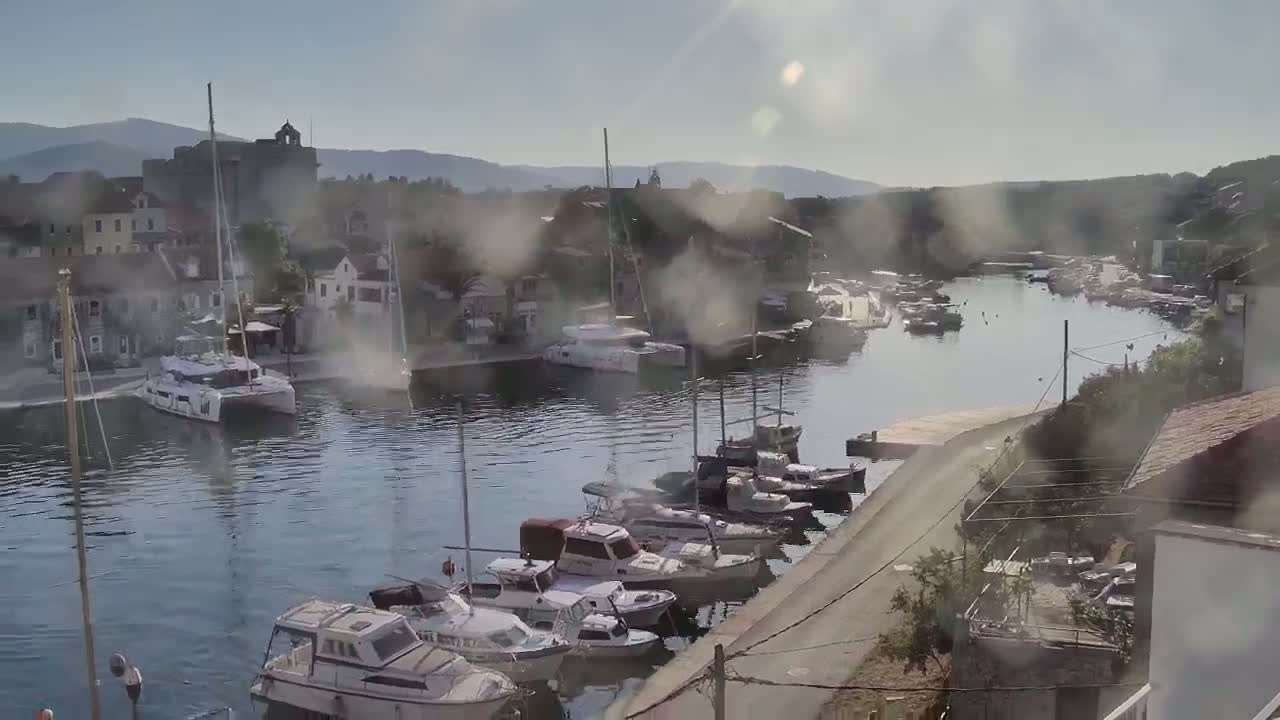 A hazy, sunny day shows a calm harbor filled with numerous boats and yachts, nestled beside a small town with stone buildings against a backdrop of distant mountains.
