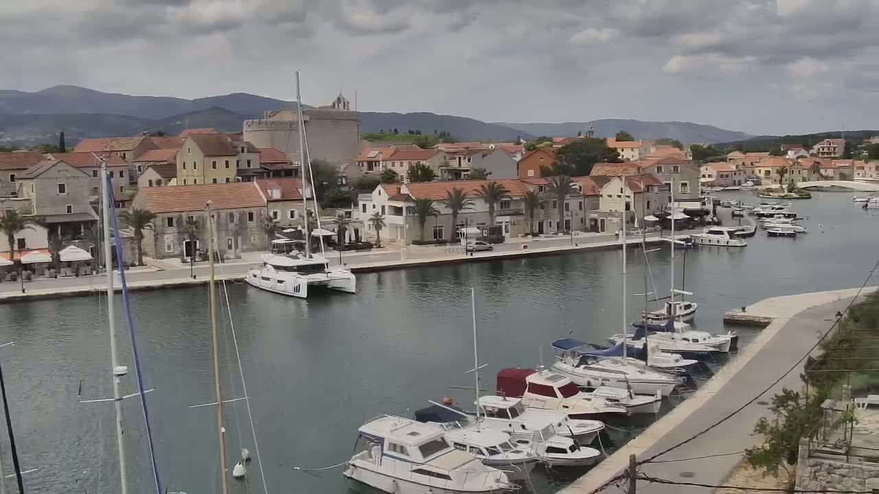 A harbor filled with sailboats and motorboats is situated in front of a stone village under a cloudy sky, backed by mountains.