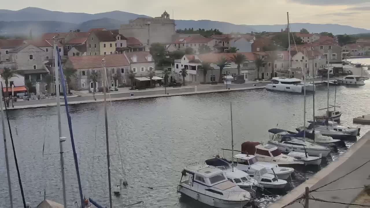 A waterfront town with stone buildings and numerous sailboats and motorboats moored in a calm harbor, under an overcast sky, with mountains visible in the background.