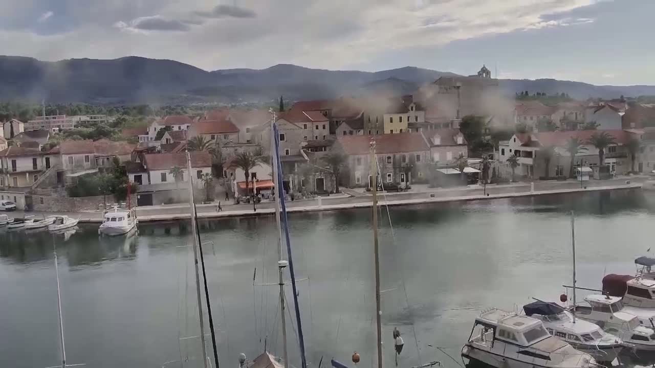 A harbor town with red-roofed buildings sits nestled against a mountain range under a cloudy sky, with numerous boats moored in the calm water.