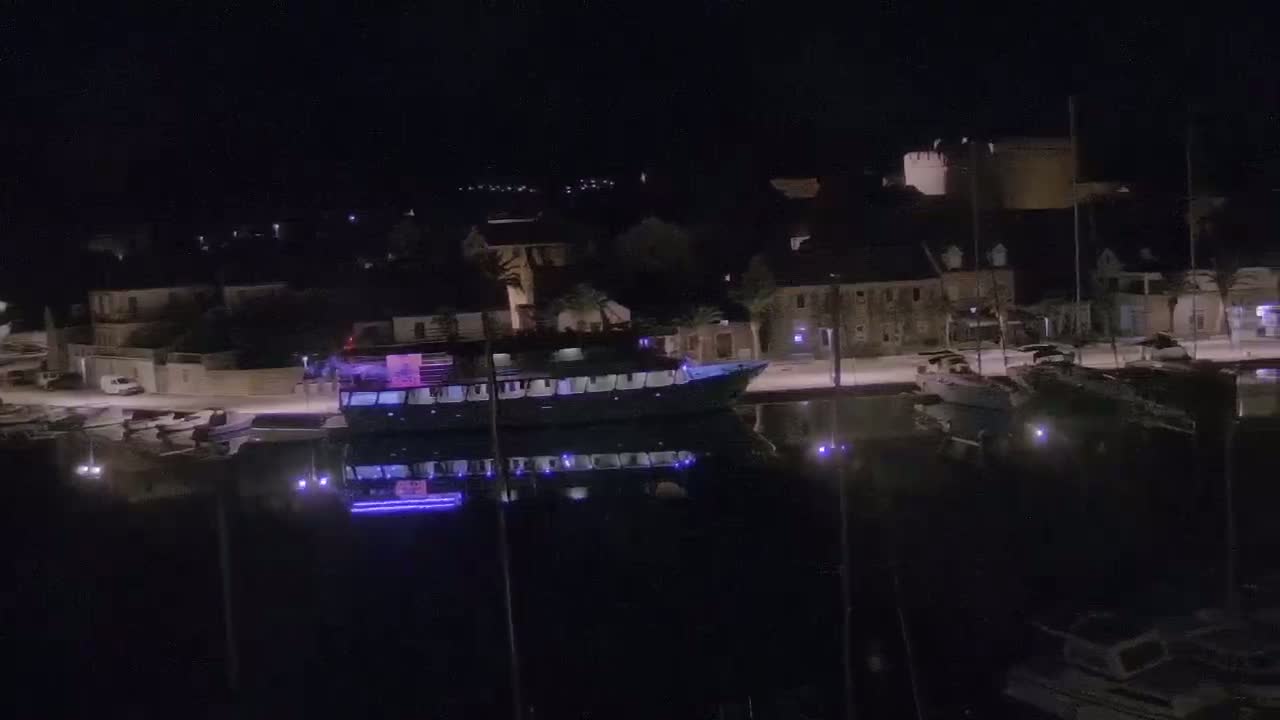 A large boat is docked at a waterfront at night, with several smaller boats nearby and buildings lining the shore under a dark sky.