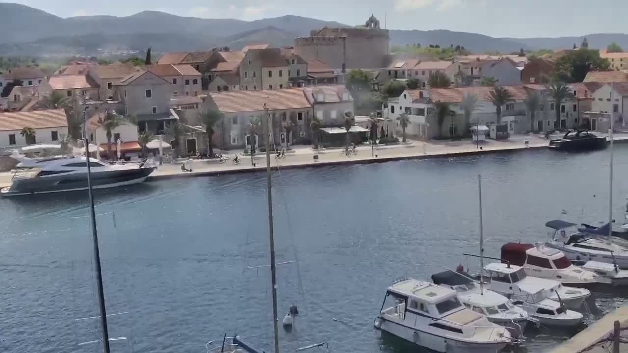 A waterfront town with stone buildings and palm trees is situated on a calm body of water where several boats and yachts are moored.