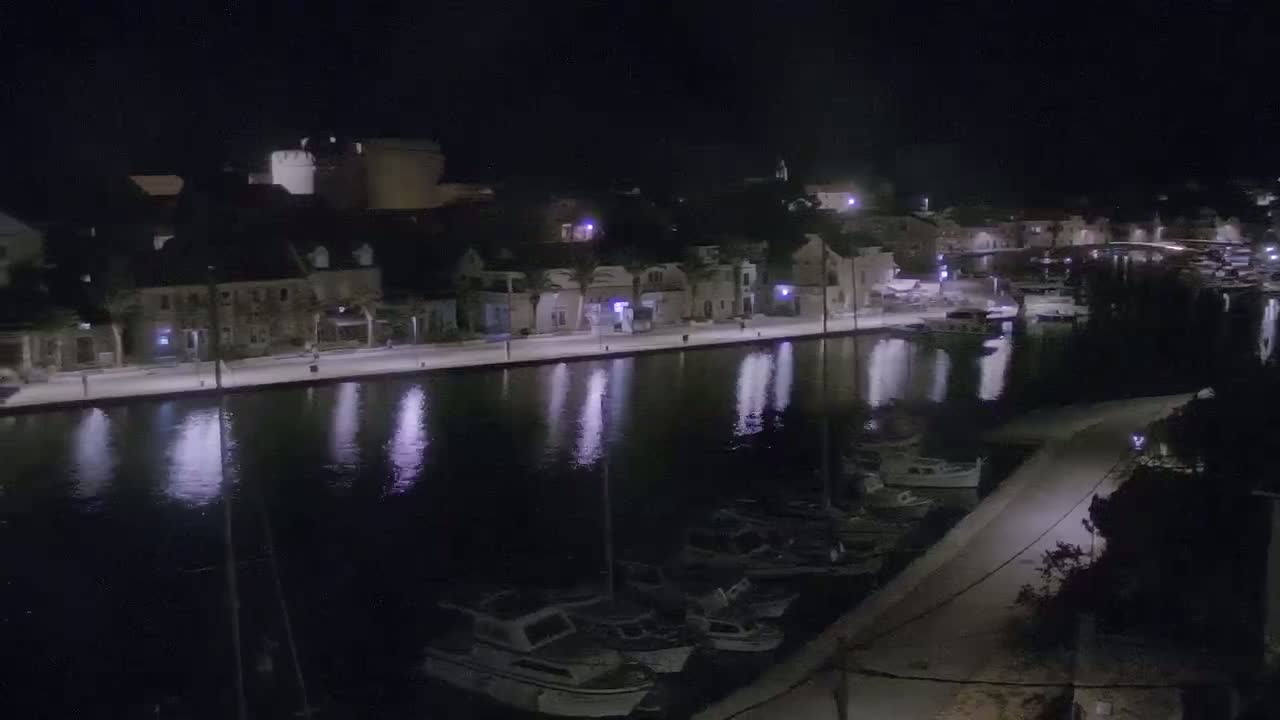 A nighttime view of a waterfront town with boats moored along a calm canal, illuminated by streetlights.