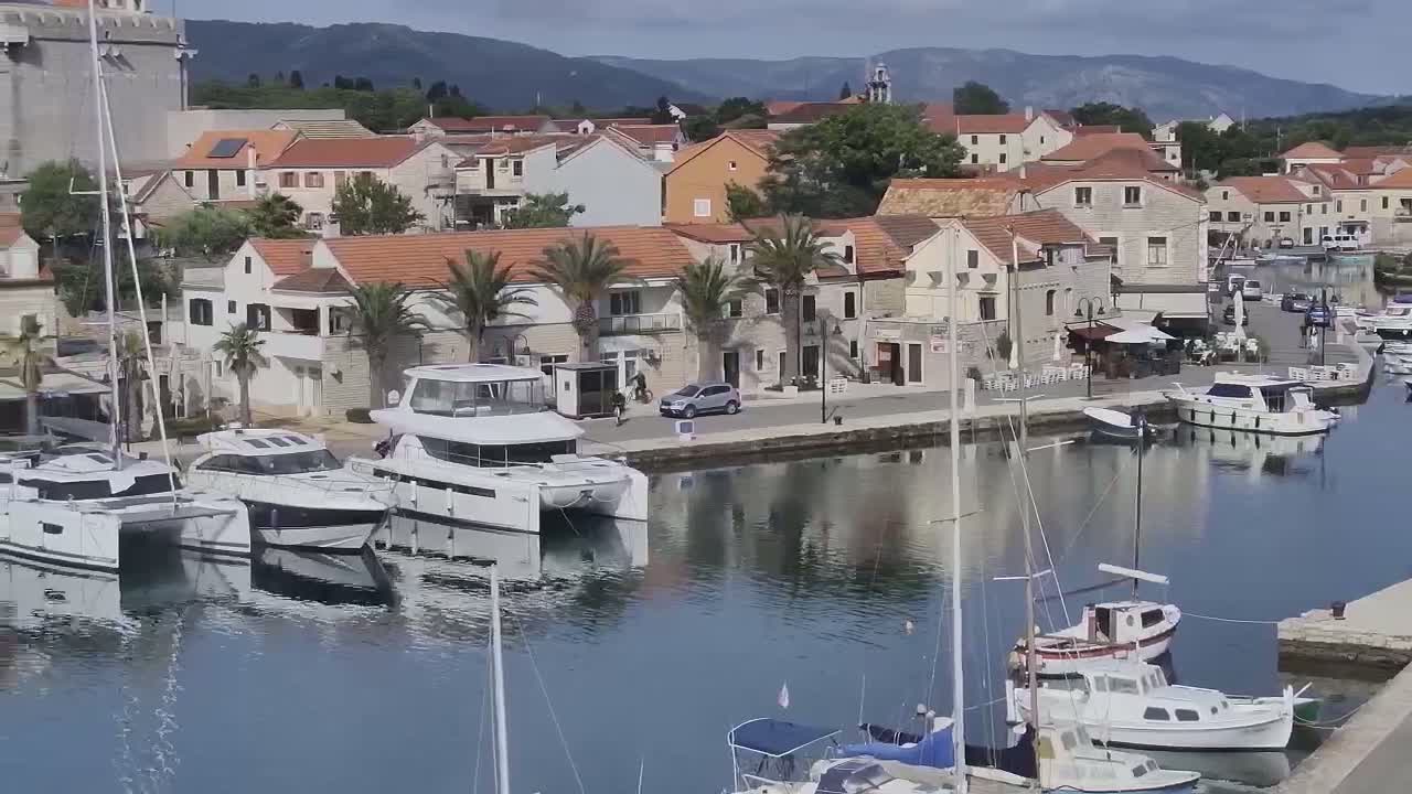 A harbor filled with sailboats and motorboats is nestled next to a village of stone buildings under a partly cloudy sky.