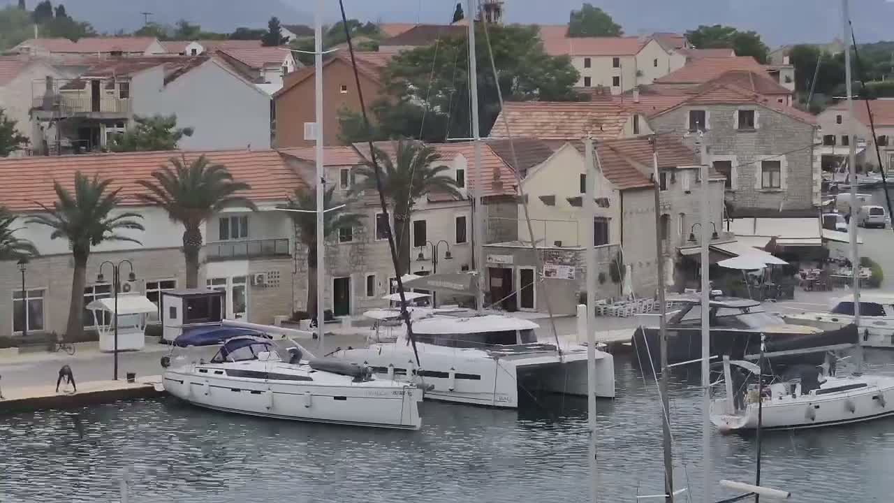 Several sailboats and motor yachts are moored in a harbor next to a stone village under an overcast sky.