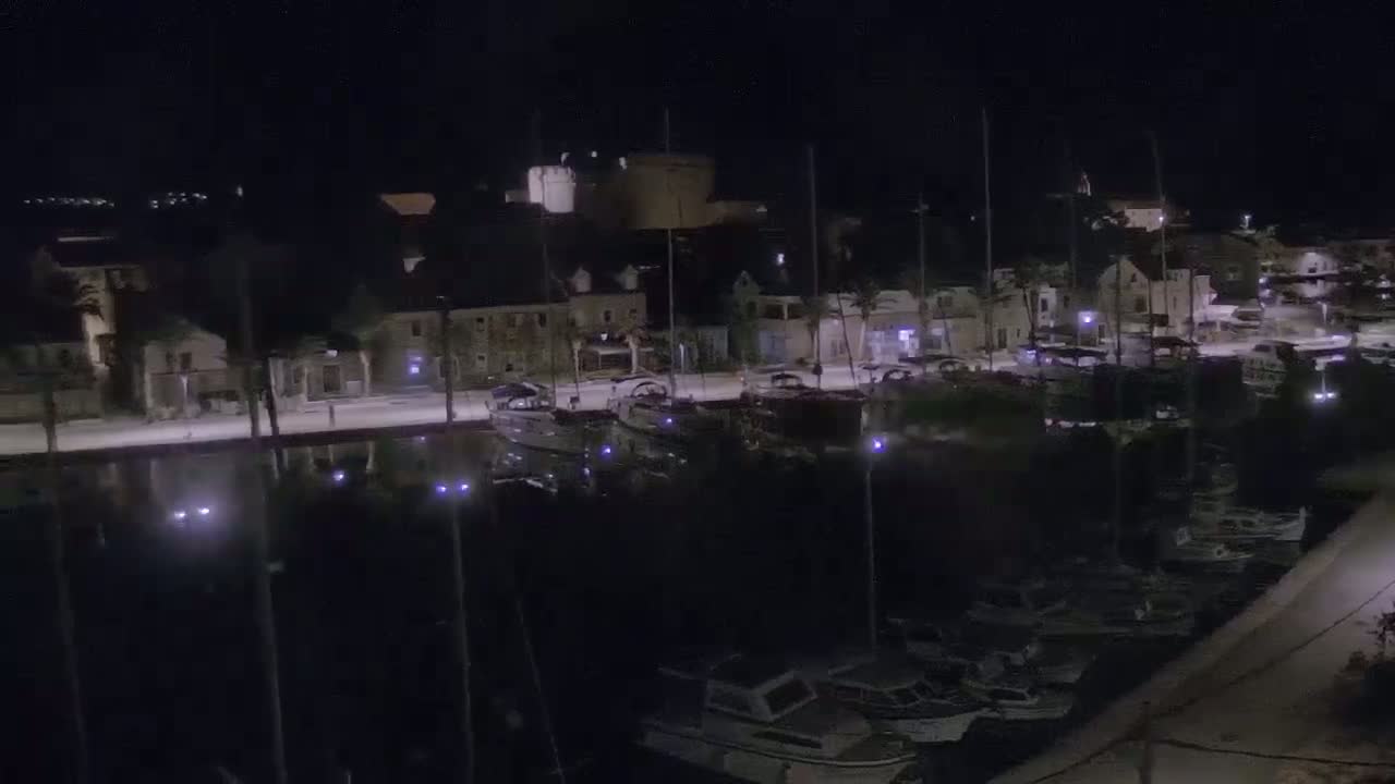 A nighttime aerial view shows a marina filled with boats docked alongside a waterfront town under a clear, dark sky.