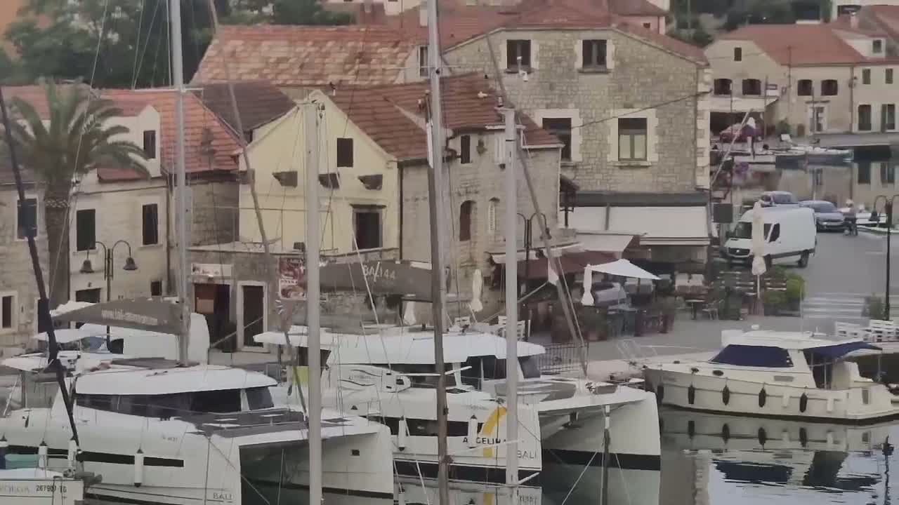 Several sailboats and motorboats are docked in a harbor next to a collection of stone buildings under an overcast sky.