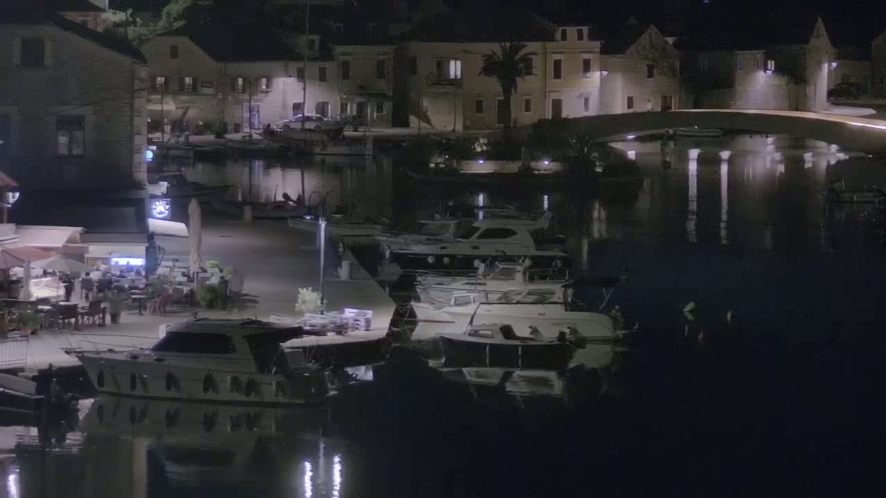 A nighttime view of a calm harbor with several boats docked along a waterfront lined with buildings and outdoor seating areas.