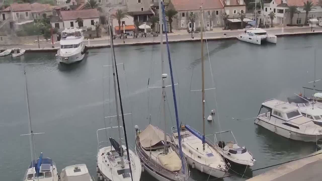 A calm harbor filled with sailboats and motorboats is viewed from above, with a town visible in the background.