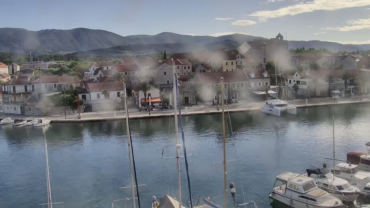 A waterfront town with low, stone buildings and palm trees sits nestled below mountains on a partly cloudy day, with numerous sailboats and motorboats moored in the calm harbor.