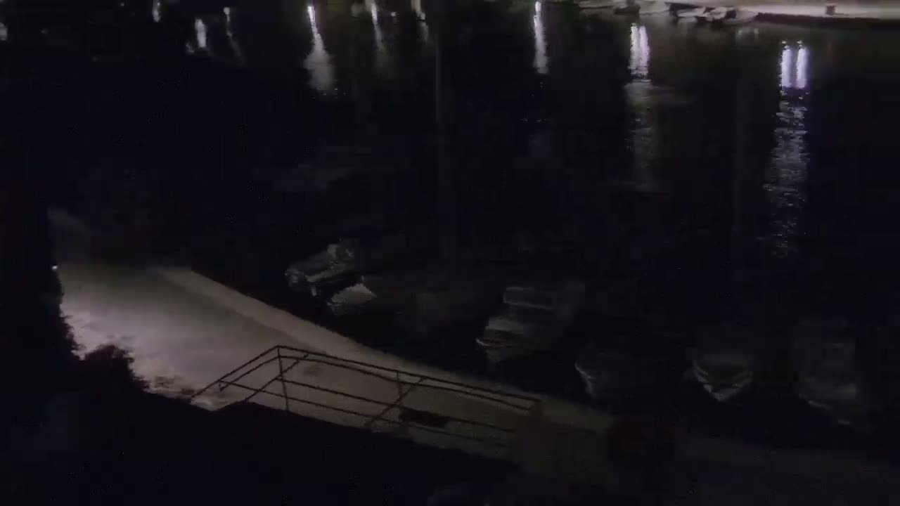 A nighttime view shows several boats moored at a dark waterfront next to a paved area with a railing.