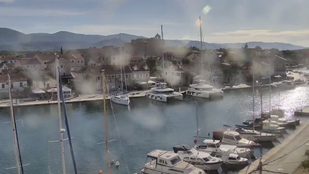 A hazy sunny day over a marina filled with sailboats and motorboats, in front of a stone village nestled against mountains.