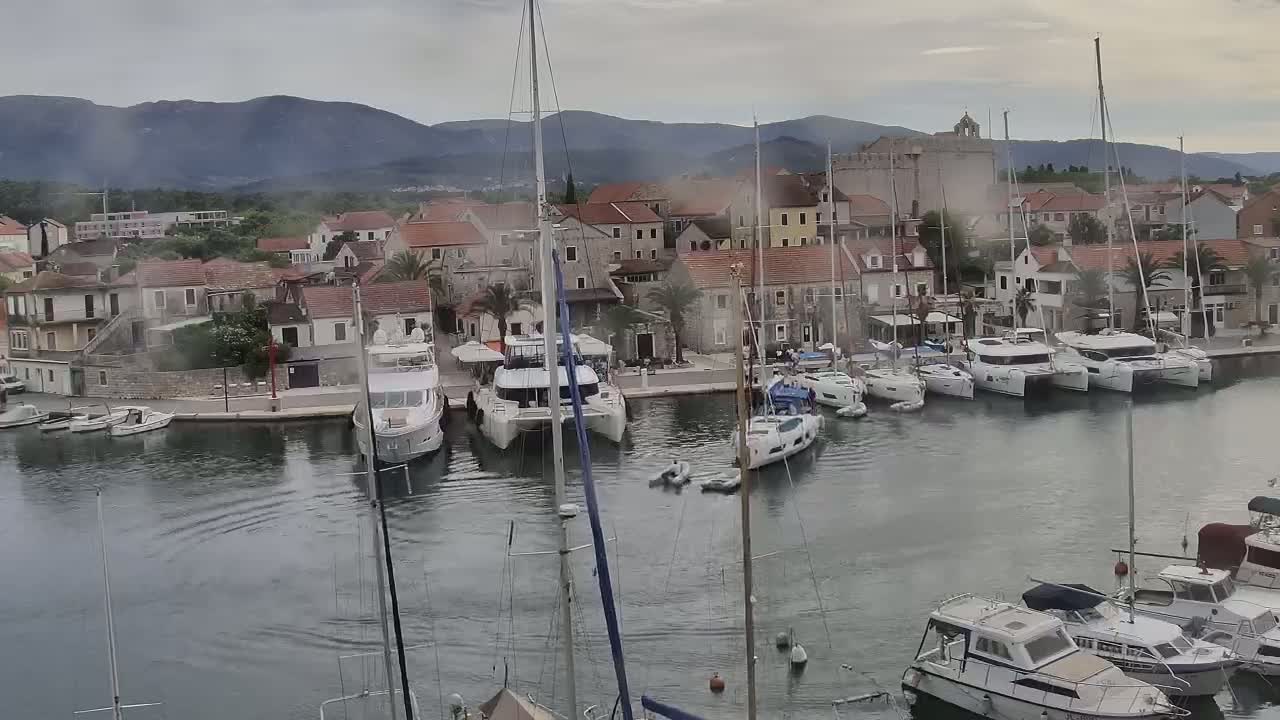 A harbor filled with yachts and sailboats is nestled next to a town of stone buildings under a cloudy sky.