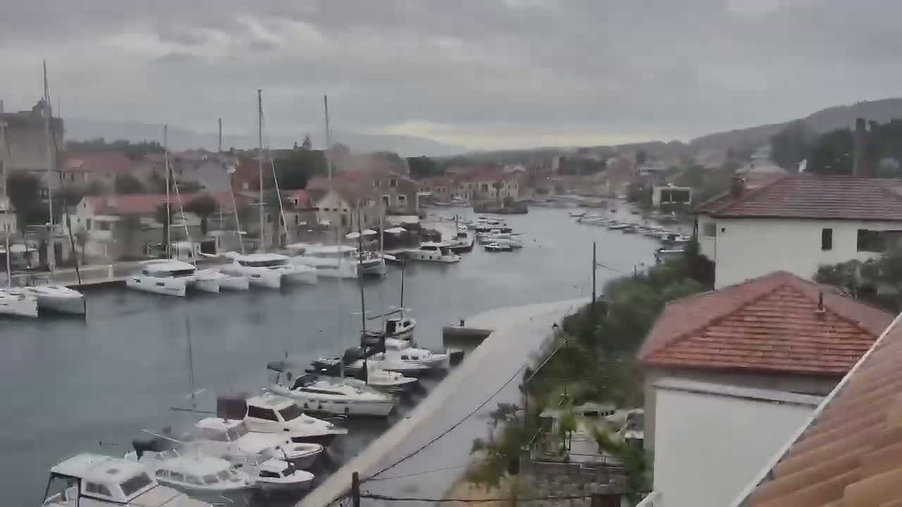 A harbor filled with sailboats and motorboats is viewed from above in the rain, with buildings and hills visible in the background.