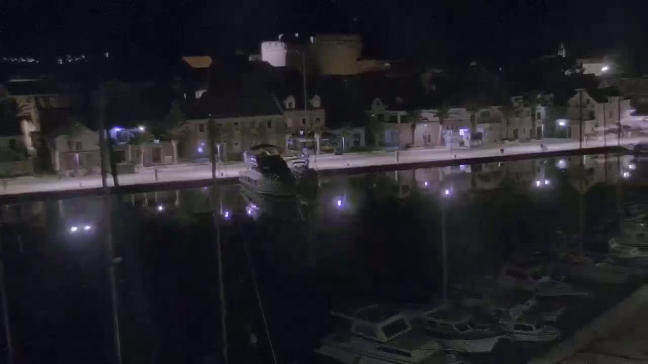 A nighttime view of a calm harbor with a docked yacht and several smaller boats, reflecting the lights of buildings lining the waterfront.