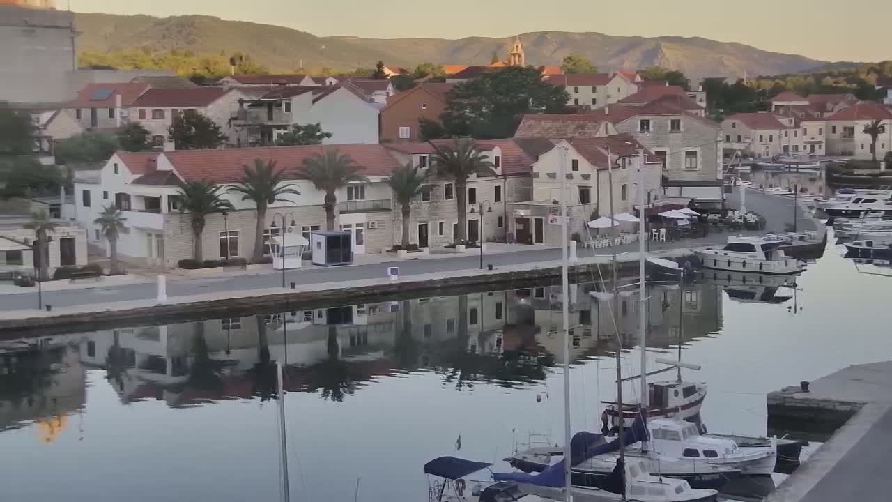 A calm harbor reflects the buildings and palm trees of a seaside town nestled against a mountain range under a clear, sunrise sky.