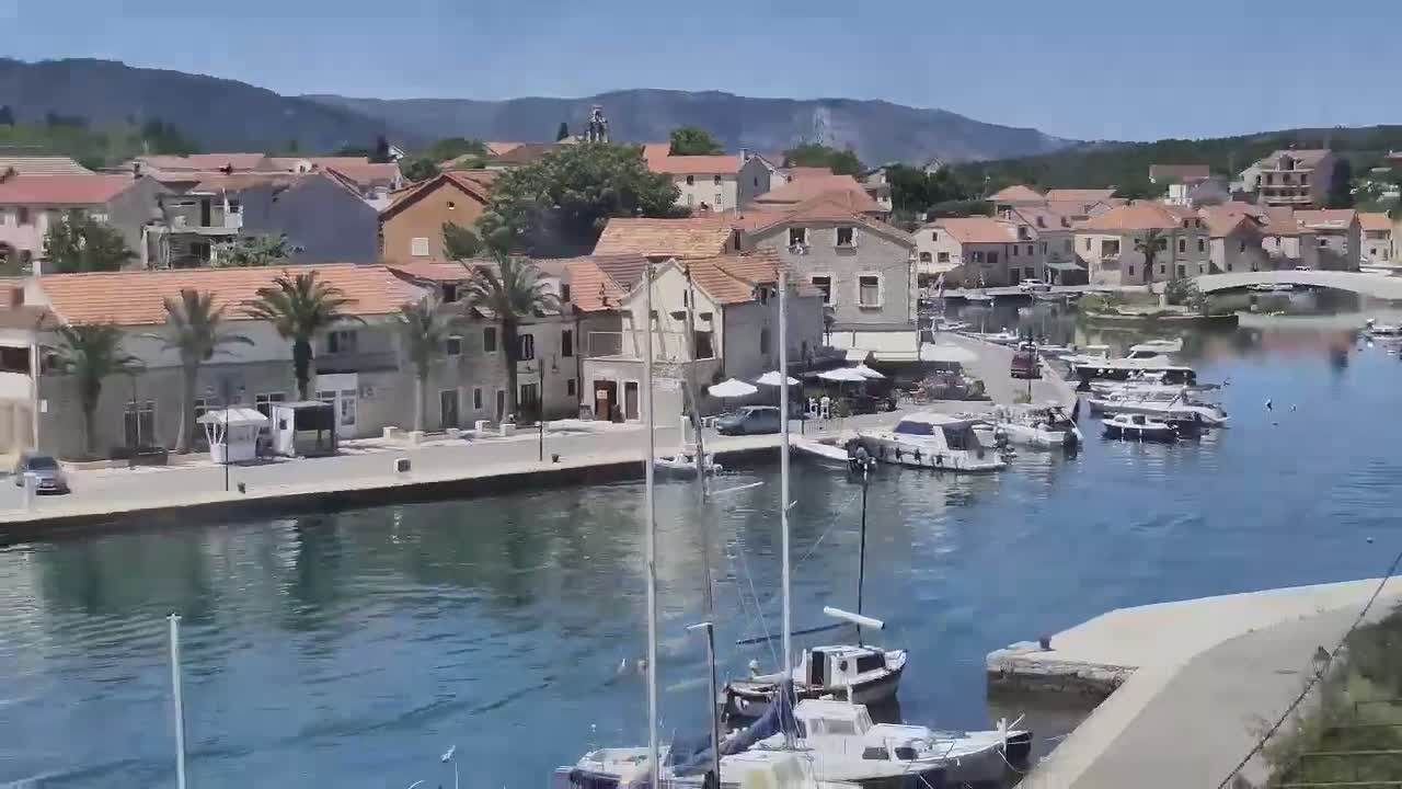 A sunny harbor town with many boats docked, surrounded by low-rise buildings and palm trees, sits nestled beneath a mountain range.