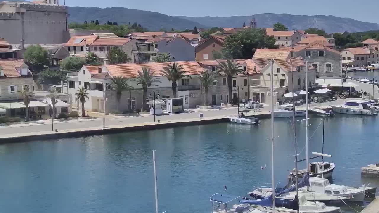 A waterfront town with Mediterranean-style buildings and palm trees, boats docked in a calm harbor, under a sunny sky.