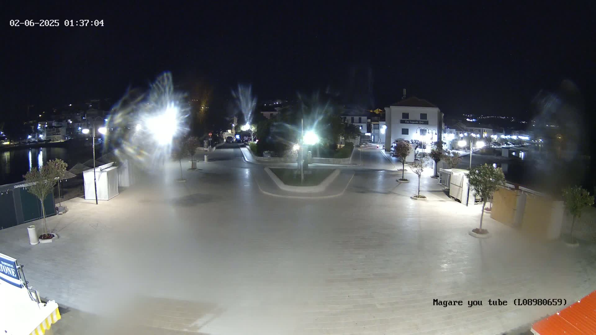 A nighttime, illuminated view of a town square near a body of water, showing several buildings and streetlights with some light fog or mist.