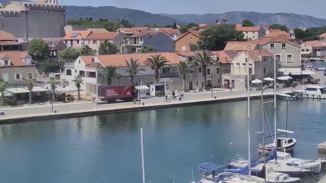 A waterfront town with red-tiled roofs, palm trees, and boats docked in a calm harbor under a sunny sky.