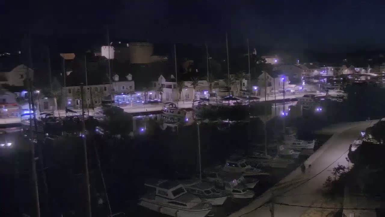 A nighttime aerial view shows a harbor filled with boats, a waterfront street lined with buildings and streetlights, under a dark sky.