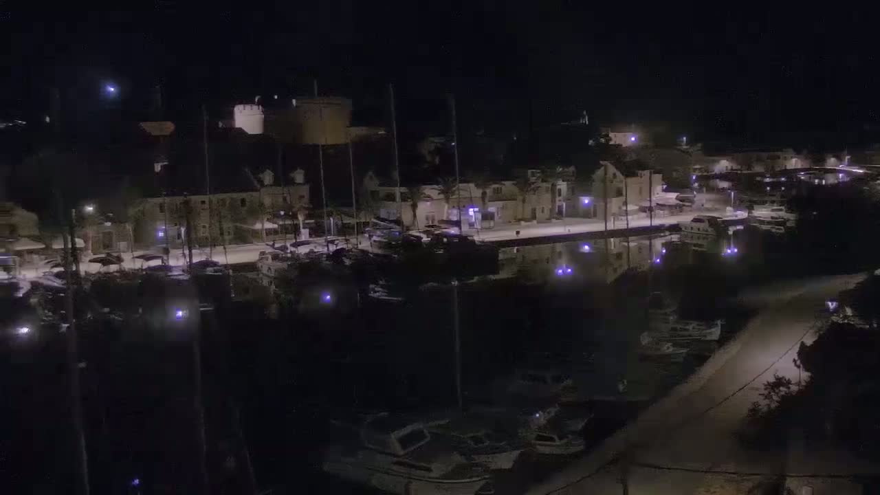 A nighttime aerial view shows a harbor filled with boats moored alongside a waterfront town under a clear, dark sky.