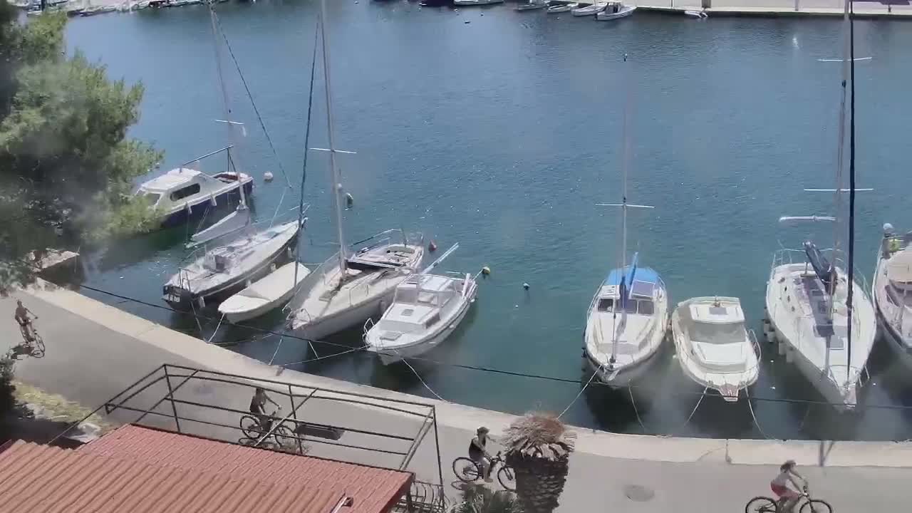 Several sailboats and motorboats are moored in a calm harbor, with people riding bicycles along a paved walkway next to the water.