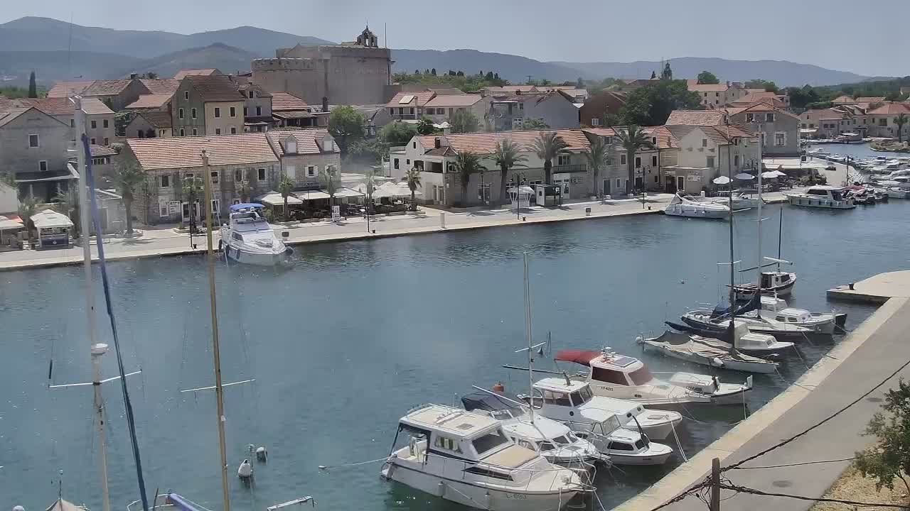 A harbor filled with numerous boats and yachts is situated beside a town of stone buildings under a clear, sunny sky, with mountains in the background.