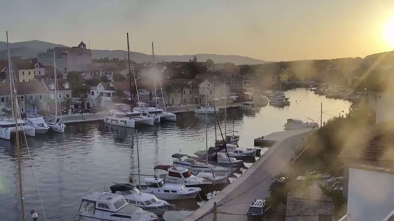 Numerous sailboats and motorboats are moored in a calm harbor at sunset, with a town and hills visible in the background.