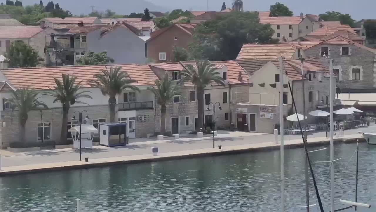 A waterfront village with stone buildings, palm trees, and outdoor seating areas is seen on a sunny day.