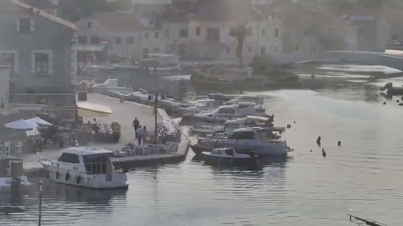 Several boats are moored along a waterfront next to buildings and a walking area, on a hazy day.