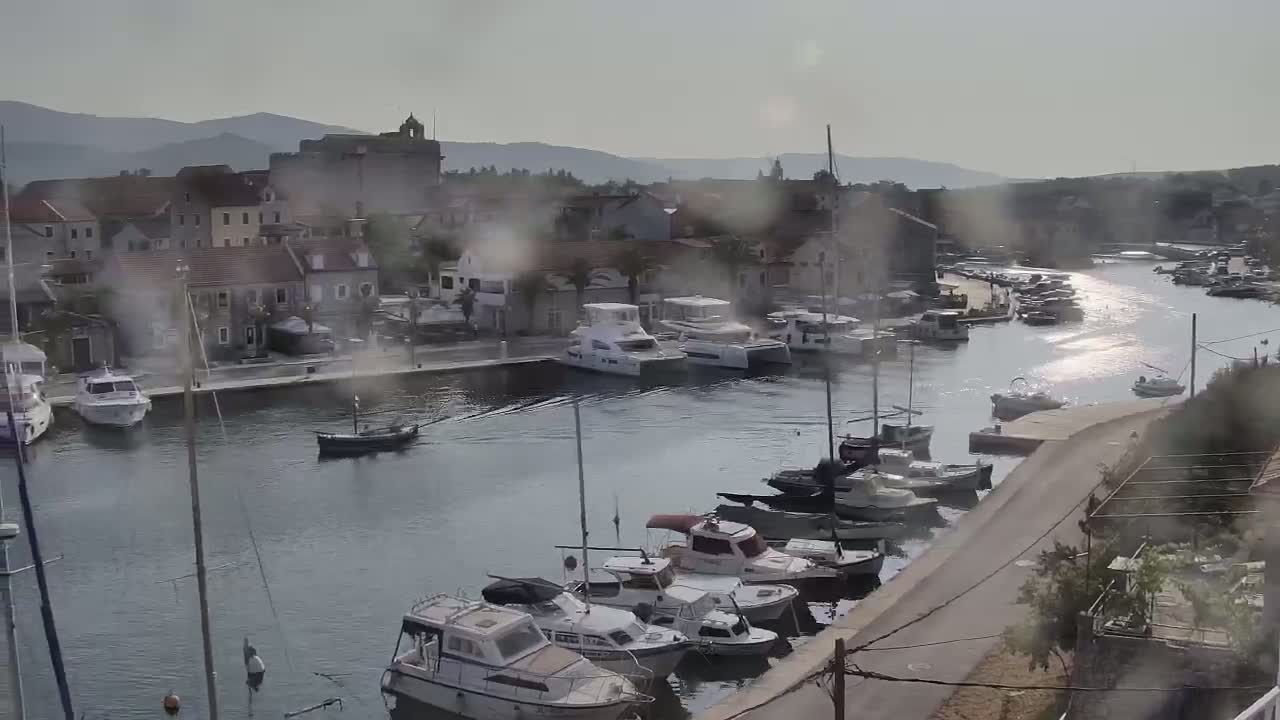 A harbor filled with numerous boats of various sizes is situated in front of a town with stone buildings under an overcast sky.