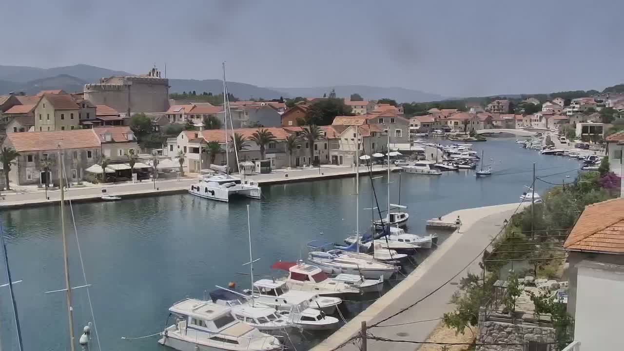 A calm harbor filled with sailboats and motorboats is nestled beside a town of low, stone buildings under a mostly clear sky.