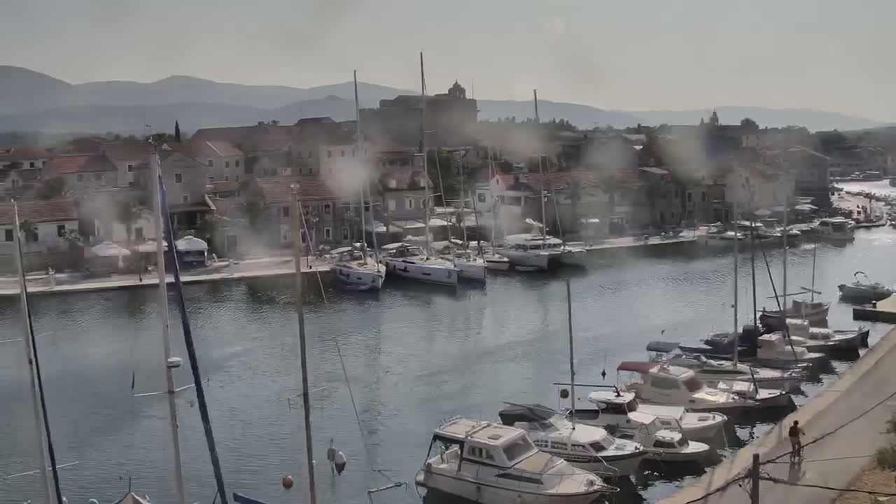 A hazy harbor filled with sailboats and motorboats is nestled beside a stone village backed by distant mountains.