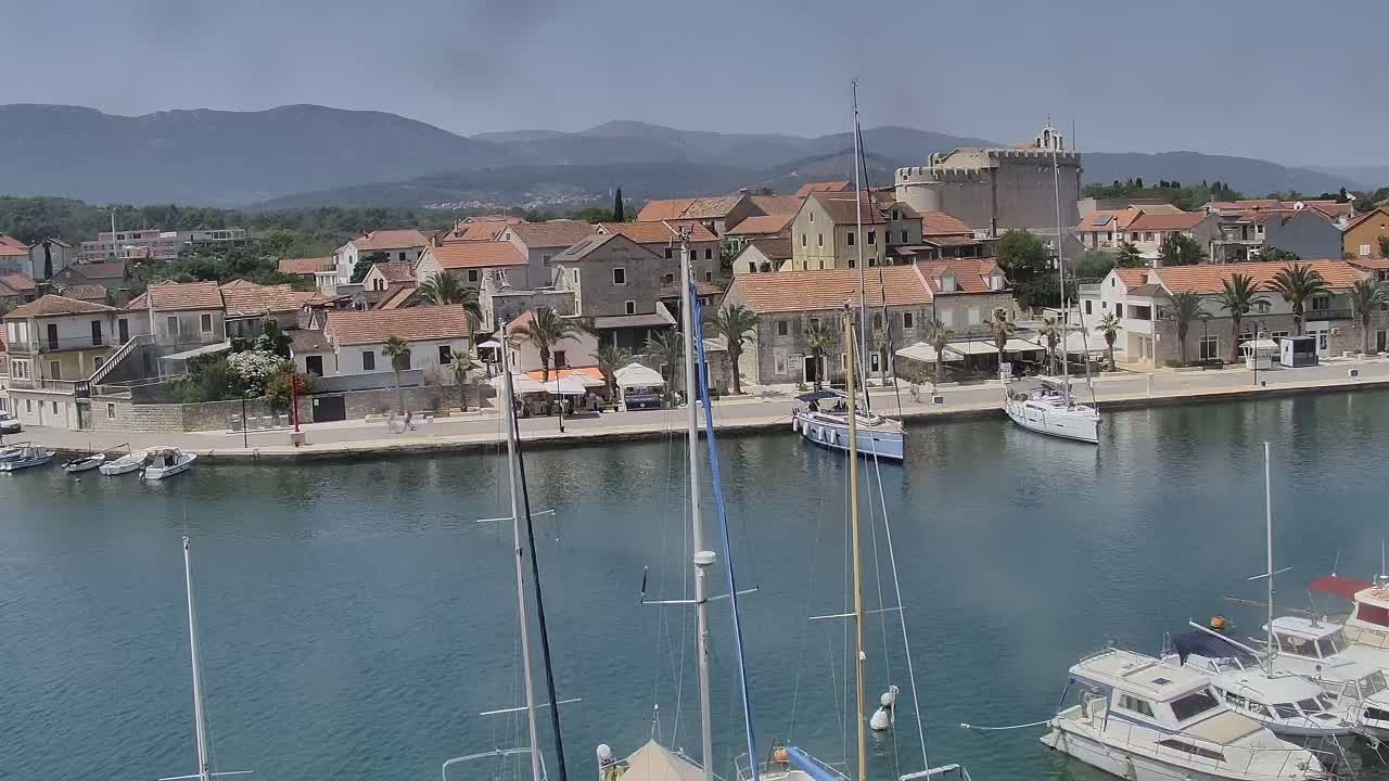 A calm harbor filled with sailboats and motorboats is situated in front of a town with red-tiled roofs, palm trees, and a stone fortress, all under a clear, sunny sky with mountains in the background.