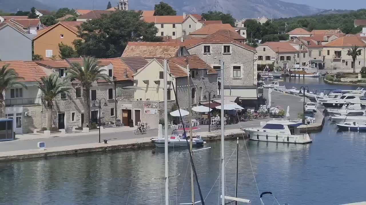 A waterfront town with stone buildings, palm trees, outdoor seating areas, and a marina filled with boats under sunny skies.
