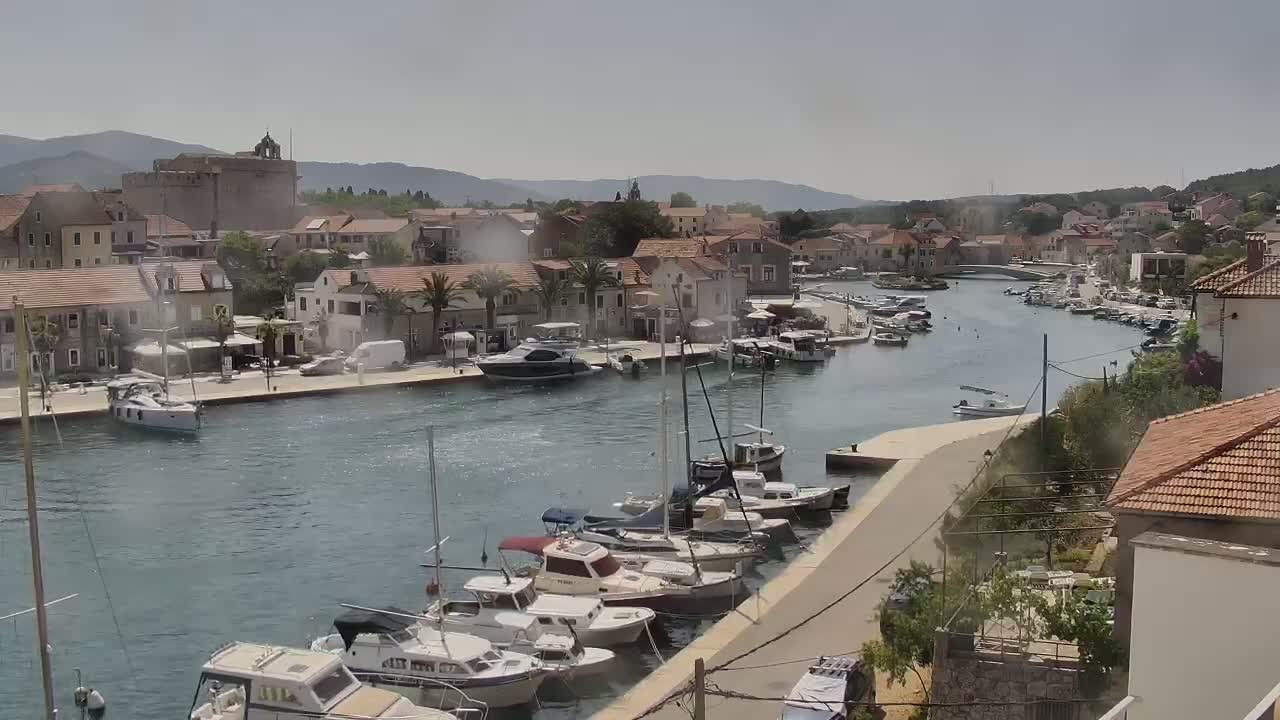 A harbor filled with boats is situated in front of a town nestled between hills on a sunny day.