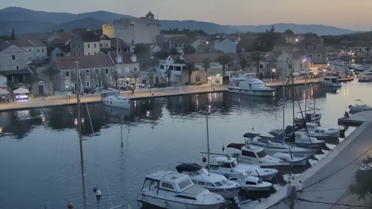 A harbor filled with boats is situated in front of a town nestled between hills on a sunny day.