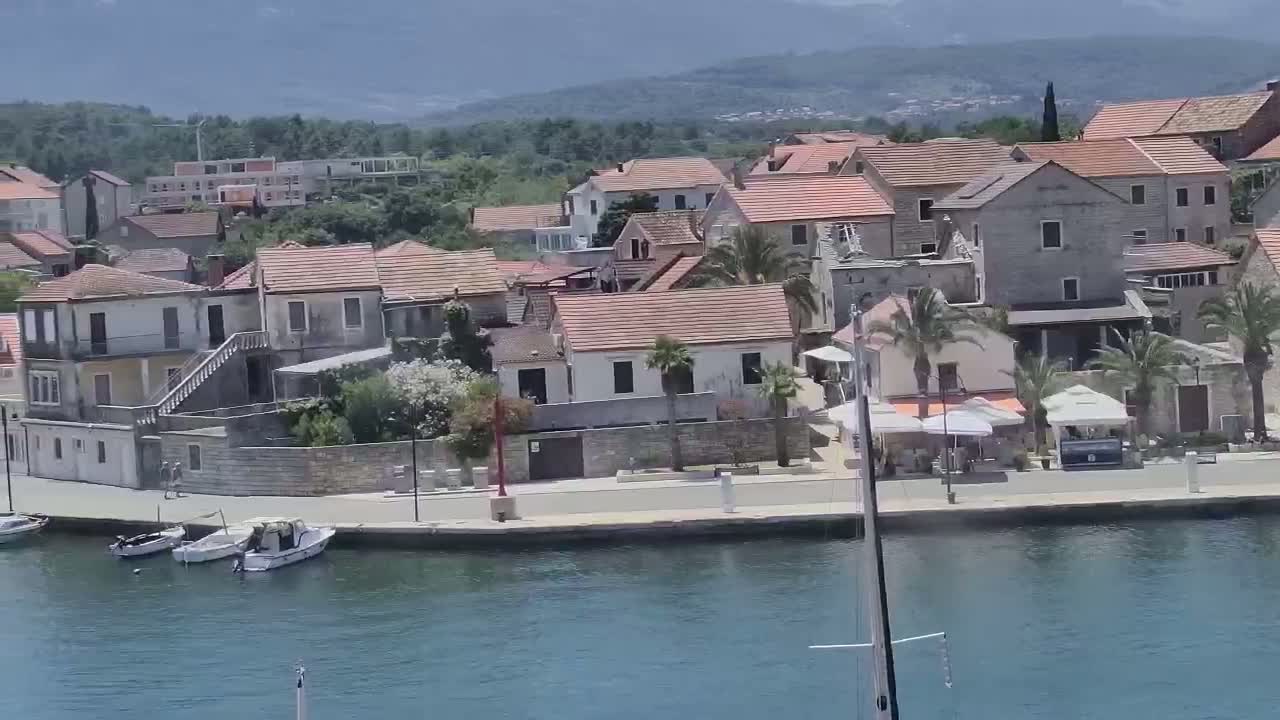 A waterfront village of closely clustered, low-rise buildings with red-tiled roofs, palm trees, and several small boats in calm water on a sunny day.