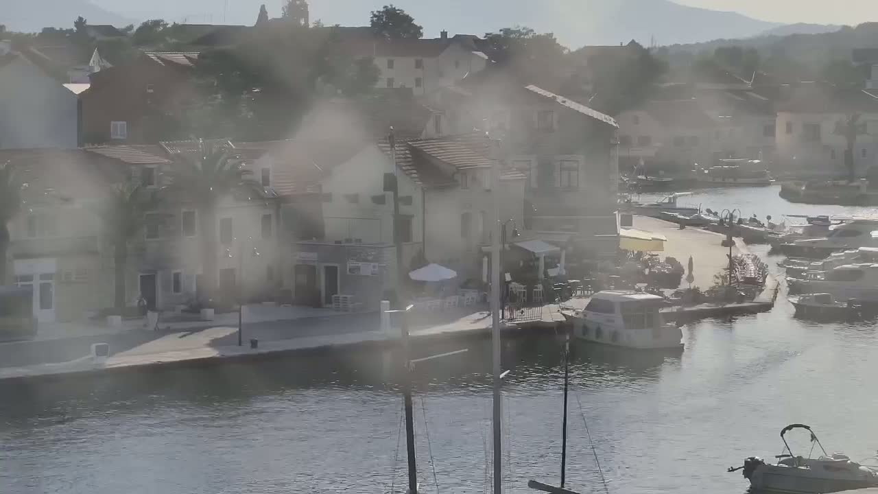 A hazy view shows a waterfront town with numerous buildings, boats docked in a calm waterway, and a few people near outdoor seating.