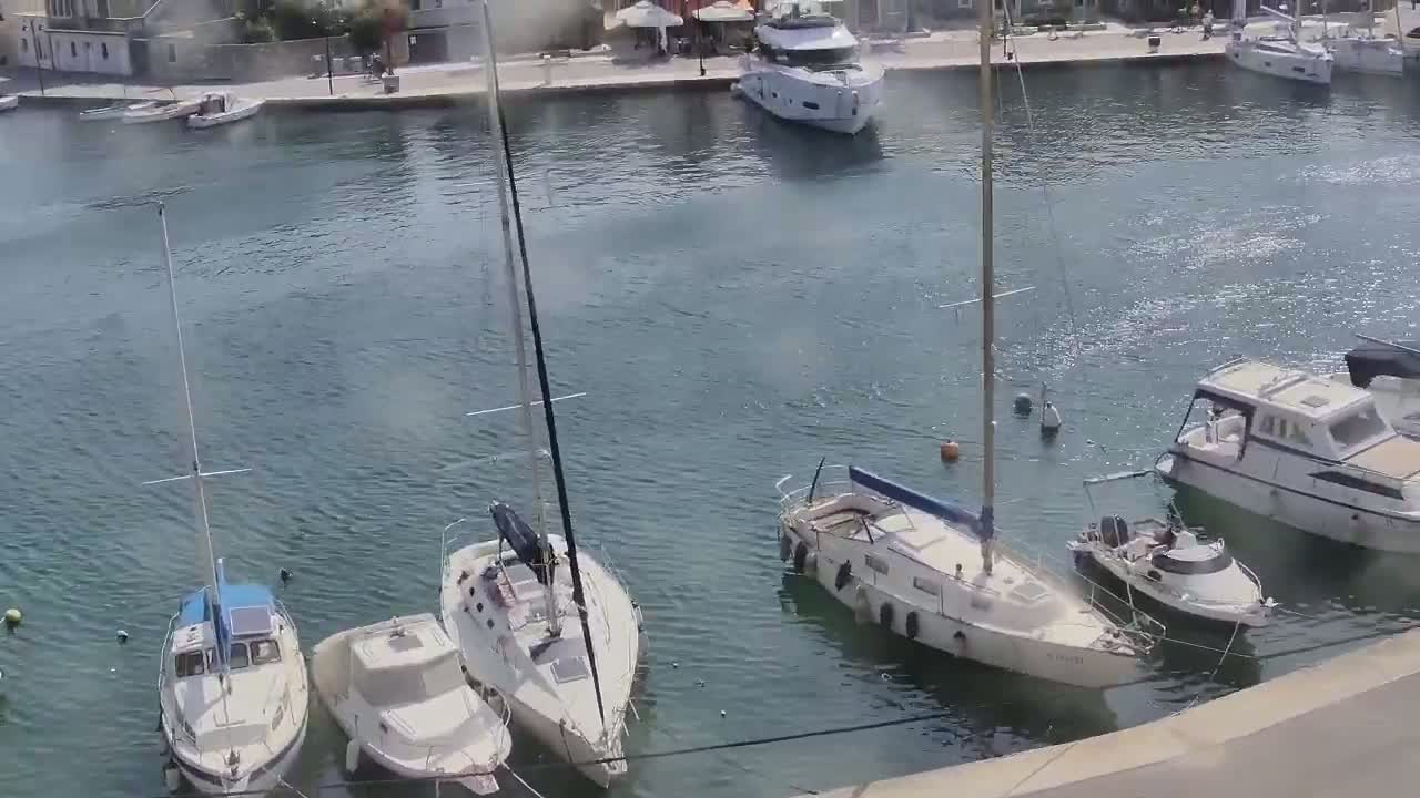 Several sailboats and motorboats are moored in a calm harbor next to a waterfront.