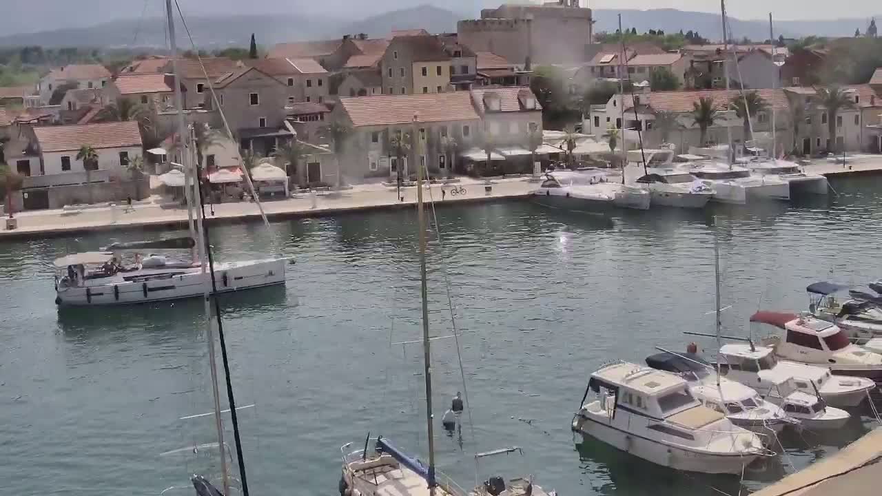 A harbor filled with sailboats and motorboats is situated next to a town of stone buildings under a partly cloudy sky.