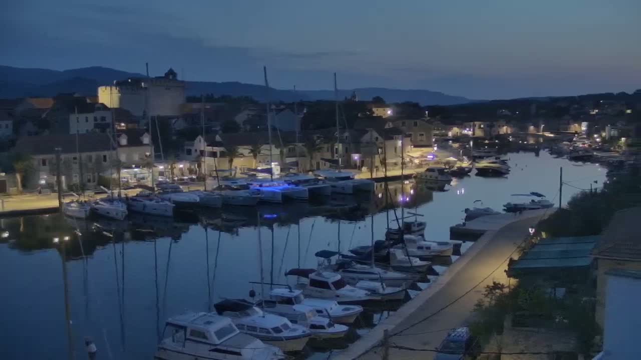Many sailboats are docked in a calm harbor at twilight, next to a waterfront town nestled against hills.