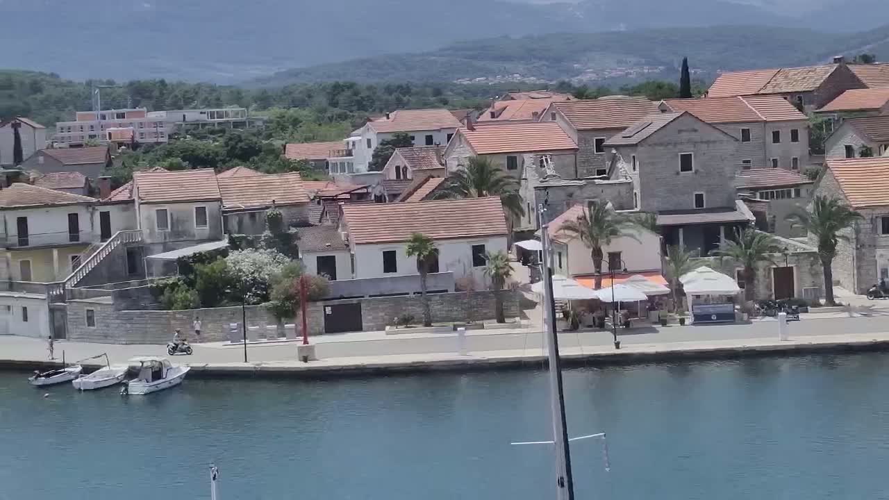 A waterfront village of low, red-tiled buildings sits under a mostly clear sky, with several small boats moored at the water's edge.