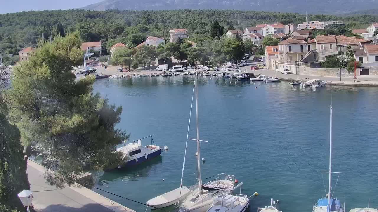 A calm harbor filled with sailboats and motorboats, nestled between a village of low-rise buildings and a mountainous background under a clear sky.