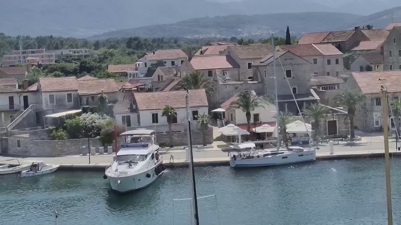 A marina with several boats, including a motor yacht and a sailboat, is situated next to a village of low, red-roofed buildings under a clear sky.